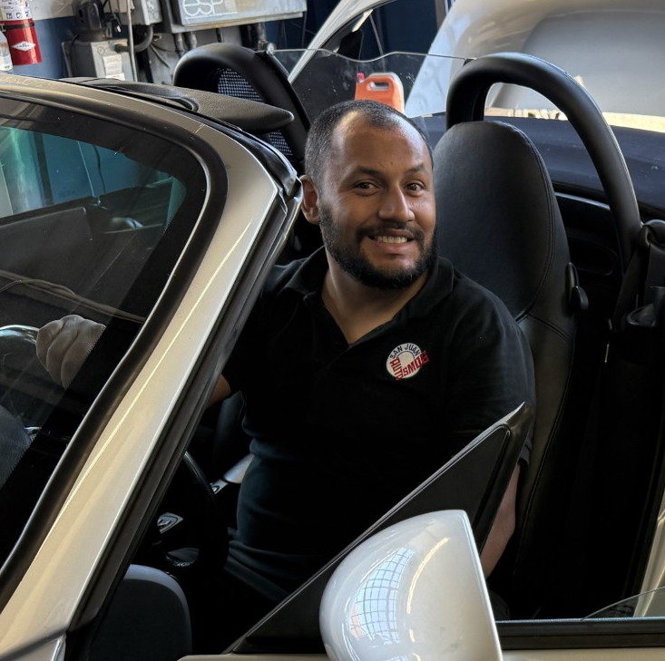 Smiling man seated in a black sports car interior, with the door open in a garage.